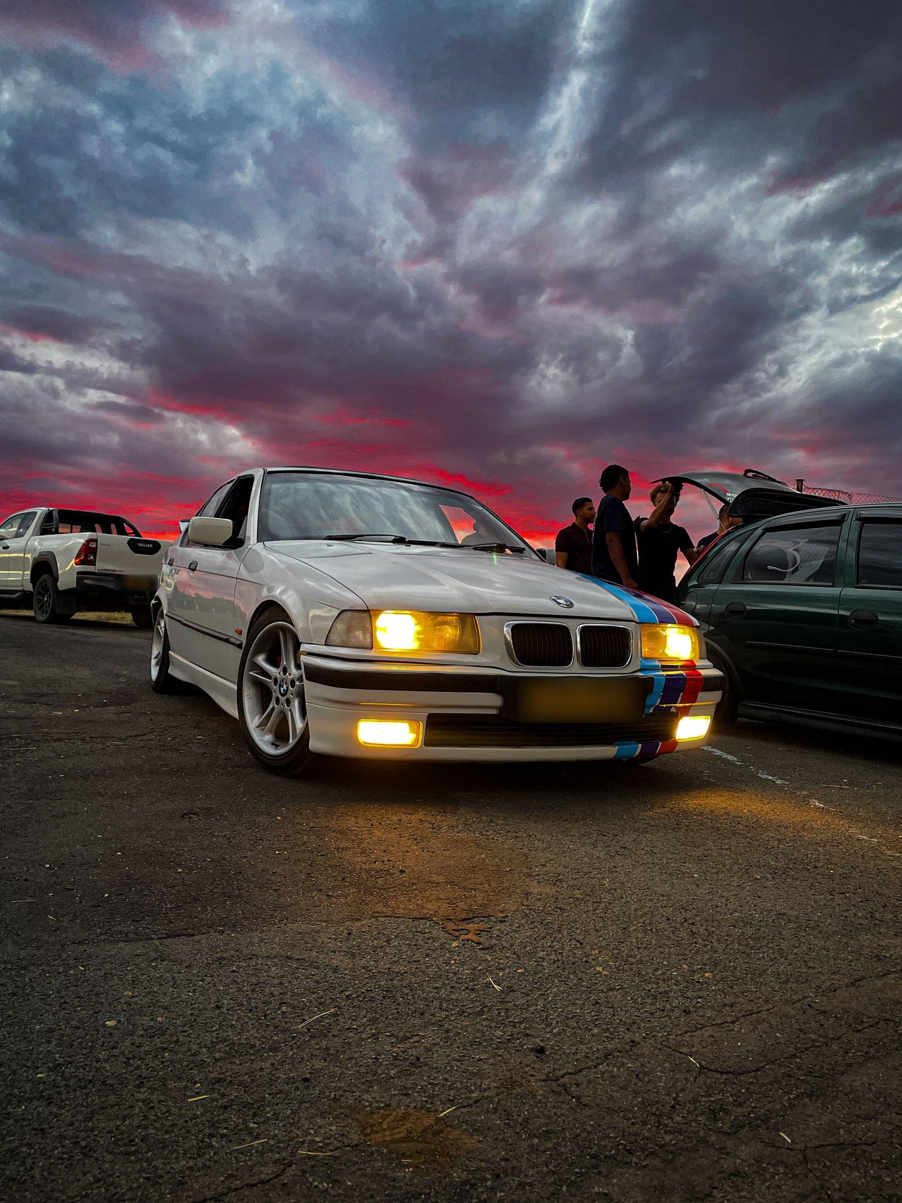 White BMW E36 with M-stripe livery, headlights on, under a red sunset sky at a car meet