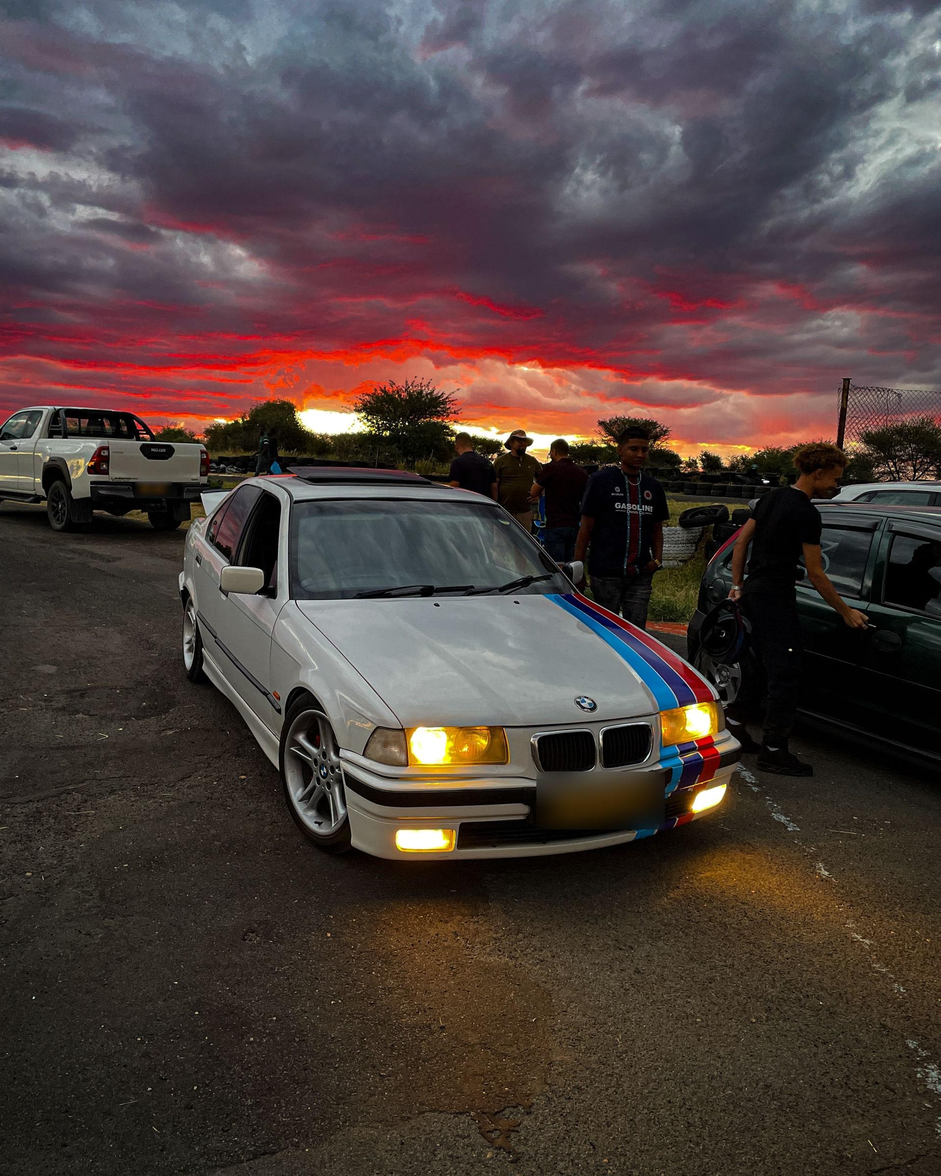 The same white BMW E36 from further back, with the sunset breaking through the clouds behind it