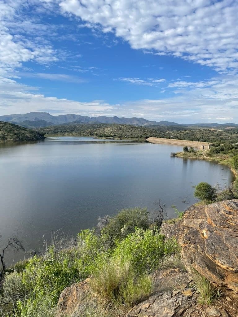 A dam ringed by green hills under a wide sky, seen from a rocky outcrop