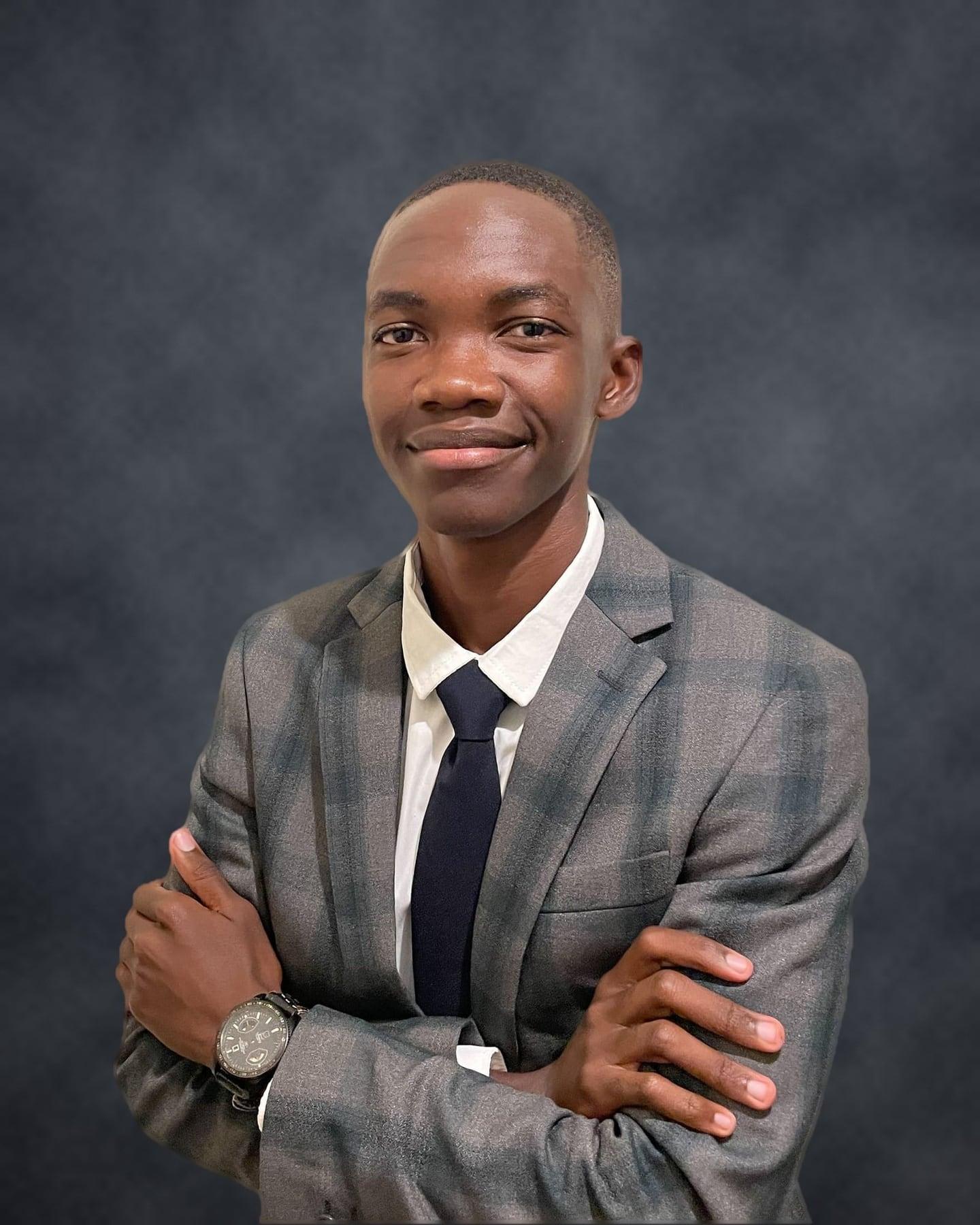 Teofilus Shaduka smiling with his arms folded, in a grey check suit and navy tie, against a dark studio backdrop